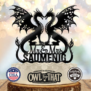 Black wedding cake topper with two winged dragons forming a heart-shaped opening, Mr and Mrs lettering, and the personalized surname SAUMENIG.