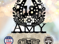 A black laser-cut 18th birthday cake topper with floral and heart details, personalized with the name 'AMY', displayed on a white-frosted cake with decorative branding seals at the bottom.