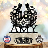 A black laser-cut 18th birthday cake topper with floral and heart details, personalized with the name 'AMY', displayed on a white-frosted cake with decorative branding seals at the bottom.