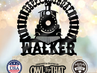 A personalized black metal cake topper shaped like a train and circular arch with the text 'HAPPY BIRTHDAY WALKER' displayed on top of a decorated cake. The background is a soft, warm bokeh light, and the image includes two badges at the bottom indicating 'Made in USA' and 'Personally Made with Love' along with the brand 'Owl Top That'.
