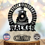 A personalized black metal cake topper shaped like a train and circular arch with the text 'HAPPY BIRTHDAY WALKER' displayed on top of a decorated cake. The background is a soft, warm bokeh light, and the image includes two badges at the bottom indicating 'Made in USA' and 'Personally Made with Love' along with the brand 'Owl Top That'.